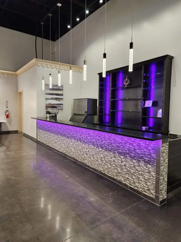 Large wet bar with sleek black granite countertops, vibrant LED underlighting, stylish tiled backsplash, built-in coolers, beer troughs, glass shelves illuminated with LED backlighting, and an ice machine for events and gatherings