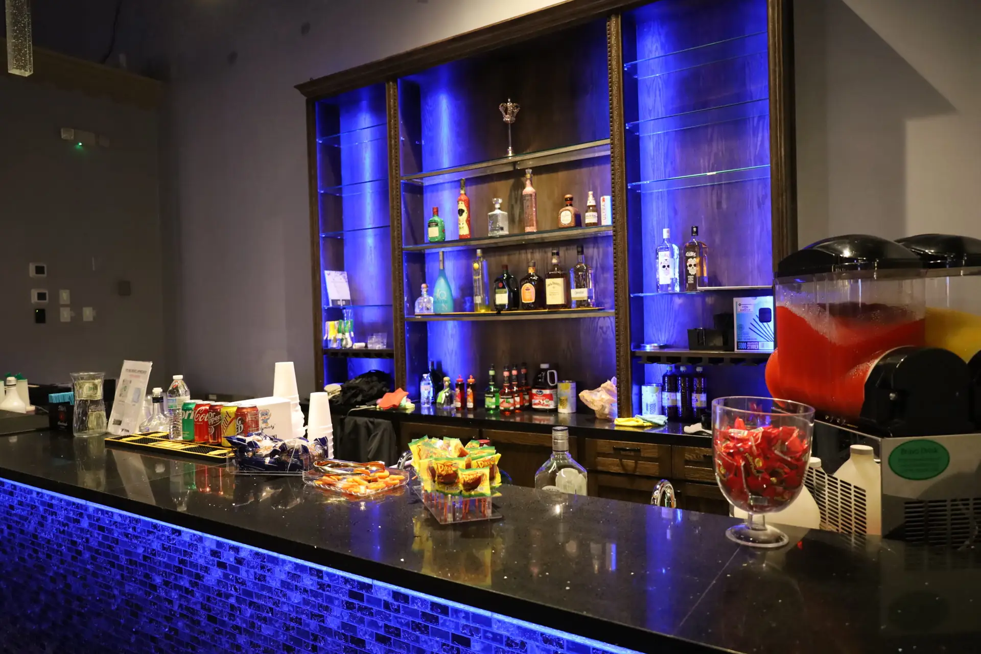 Large wet bar with sleek black granite countertops, vibrant LED underlighting, stylish tiled backsplash, built-in coolers, beer troughs, glass shelves illuminated with LED backlighting, and an ice machine for events and gatherings