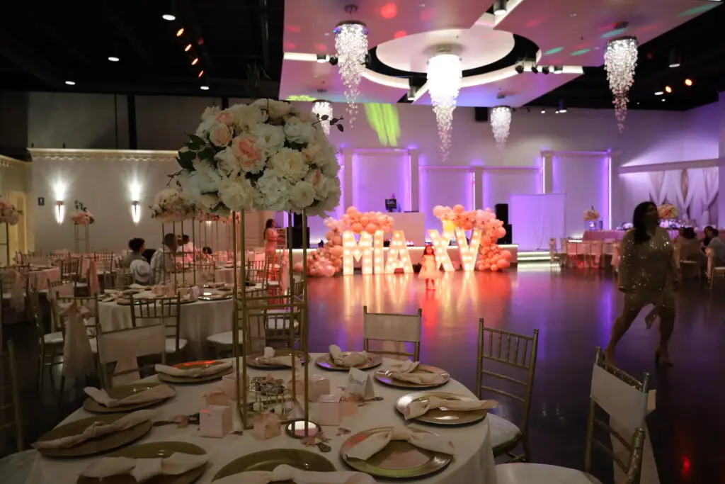 Quinceañera party in a majestic banquet hall with pink and gold decor, featuring elegant table settings, floral arrangements, and ambient lighting