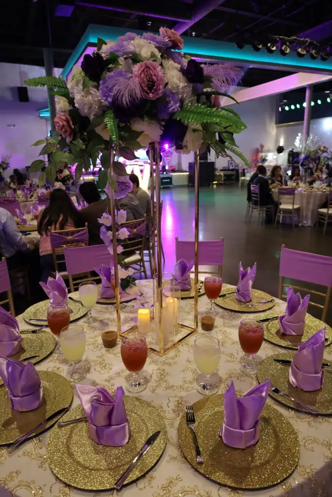 Quinceañera party in a majestic banquet hall with purple and gold decor, featuring luxurious table settings, floral arrangements, and elegant lighting