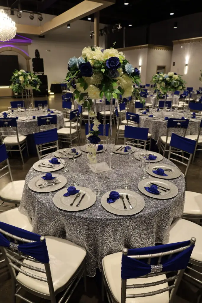 Quinceañera party in a majestic banquet hall with blue and silver decor, featuring elegant table settings, shimmering accents, and dazzling lighting