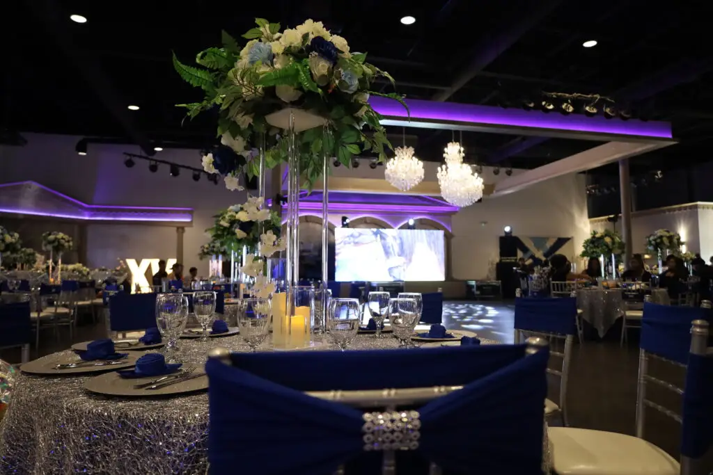 Quinceañera party in a majestic banquet hall with blue and silver decor, featuring elegant table settings, shimmering accents, and dazzling lighting