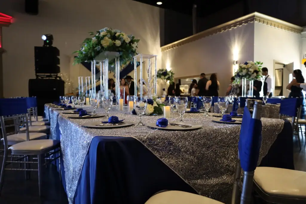 Quinceañera party in a majestic banquet hall with blue and silver decor, featuring elegant table settings, shimmering accents, and dazzling lighting